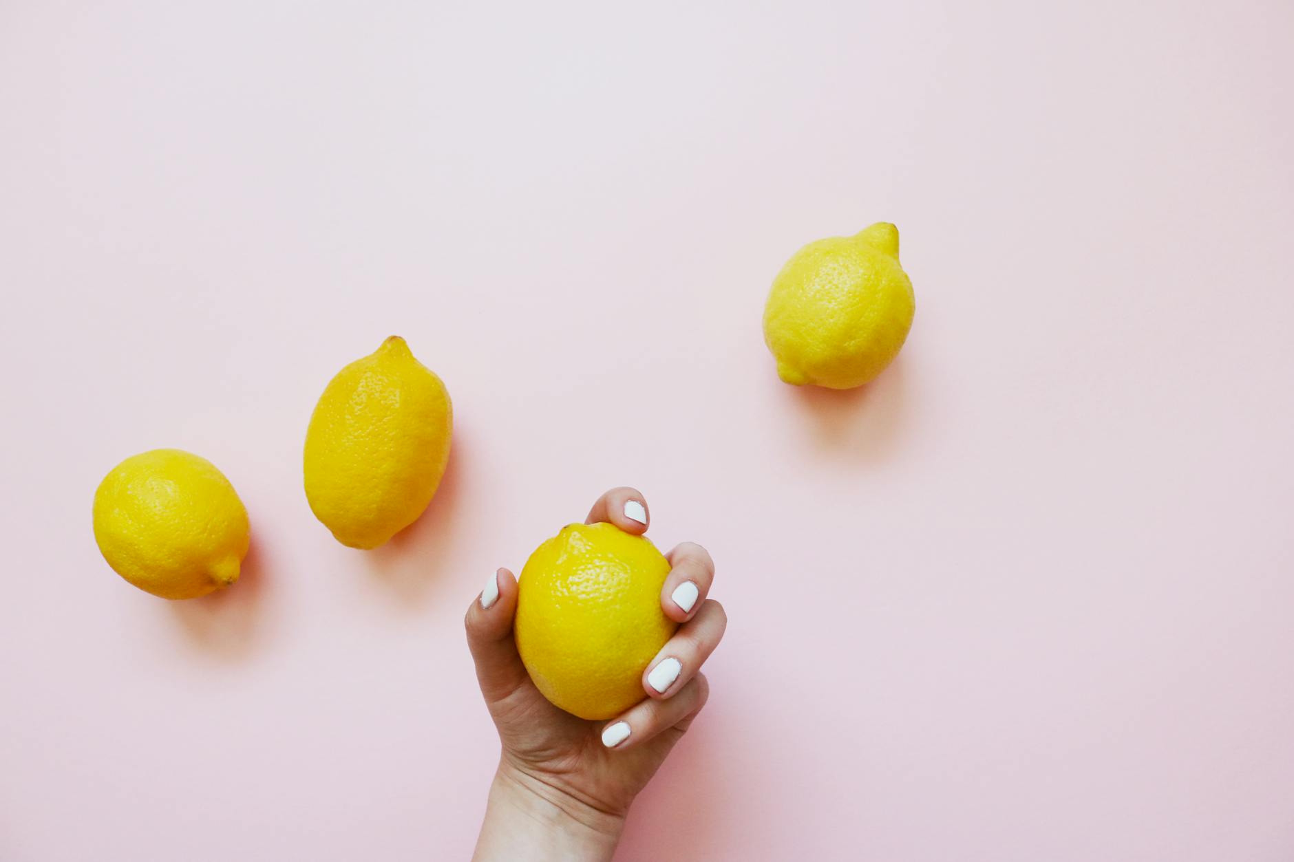 Una mano sosteniendo un limón brillante sobre un fondo rosa suave, rodeada de limones adicionales