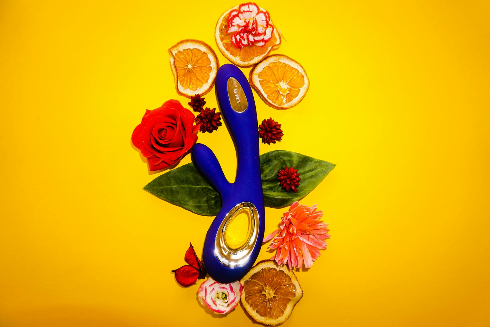 Vibrador azul rodeado de flores y rodajas de naranja sobre fondo amarillo